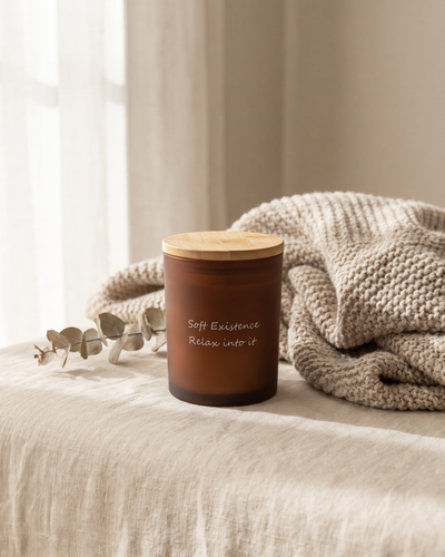 The Golden Hour soy candle in its frosted amber glass vessel with a bamboo lid, resting on linen beside a knitted throw and dried eucalyptus