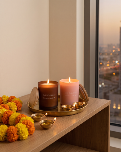 Two lit soy candles in amber and blush glass among brass diyas and a marigold garland on an oak console for Diwali