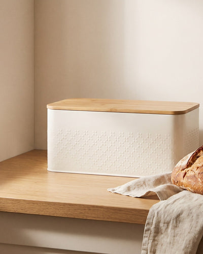 Cream steel bread bin with bamboo lid beside a fresh sourdough loaf on an oak kitchen counter