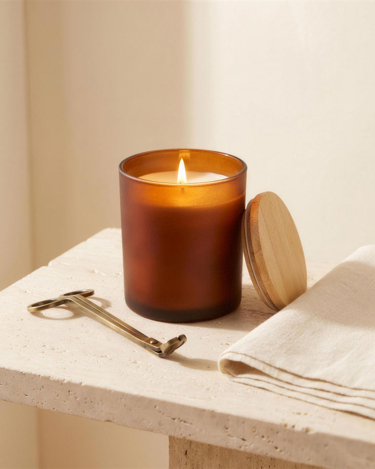 An amber glass soy candle lit beside a brass wick trimmer and folded linen on a stone surface.