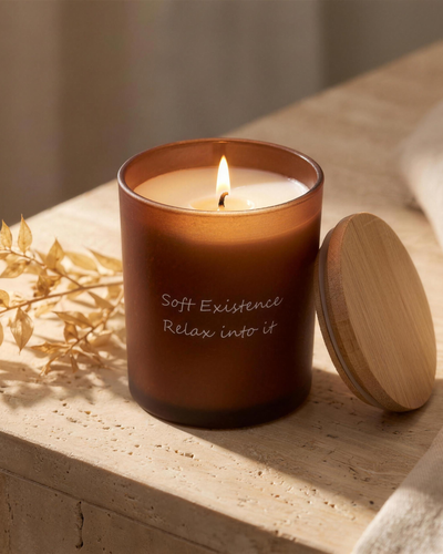 A lit Soft Existence amber glass soy candle with a clean even melt pool and bamboo lid on a travertine ledge