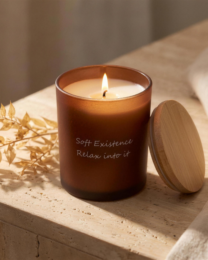 A lit Soft Existence amber glass soy candle with a clean even melt pool and bamboo lid on a travertine ledge
