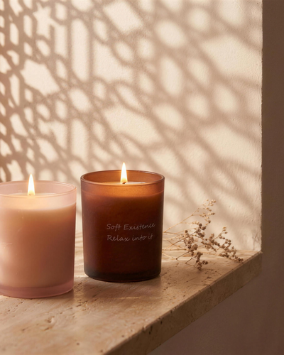 Amber and blush pink Soft Existence scented candles lit on a travertine ledge in a Dubai apartment with lattice window light