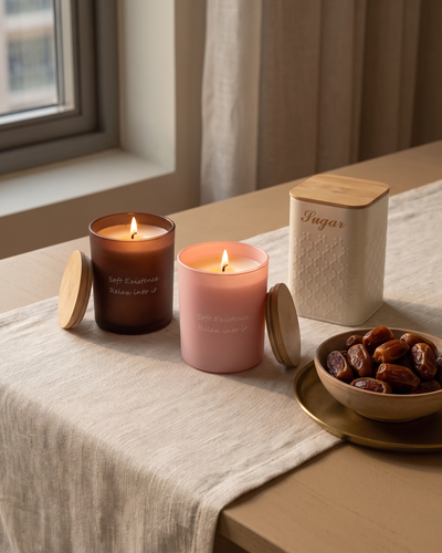 Two lit soy candles in amber and blush glass beside a cream sugar canister and a bowl of dates on a linen table runner