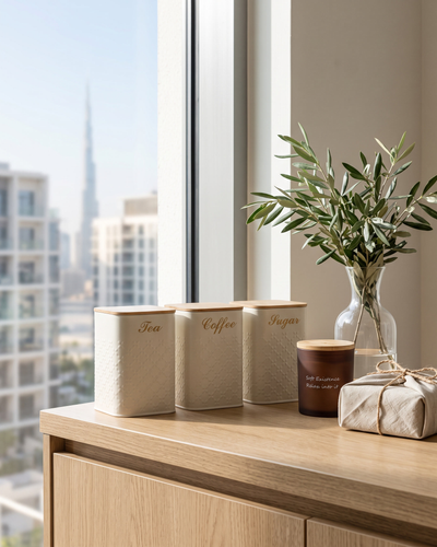 Cream tea, coffee and sugar canisters with an amber soy candle and a linen-wrapped parcel on an oak counter with the Dubai skyline behind