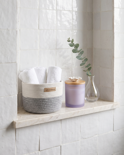 Small woven cotton basket holding rolled white towels beside a lavender soy candle on a marble bathroom shelf against white tiles