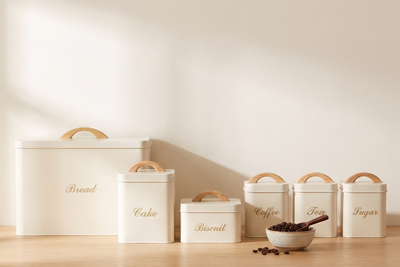 Cream six-piece kitchen canister and bread bin set with wooden arch handles lined up on an oak counter beside a bowl of coffee beans