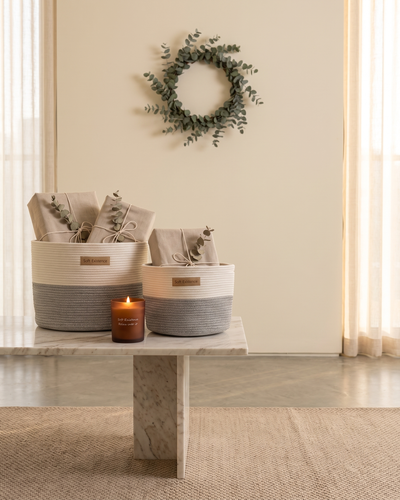 Woven cotton baskets holding linen-wrapped parcels beside a lit amber soy candle and a eucalyptus wreath for a quiet Christmas