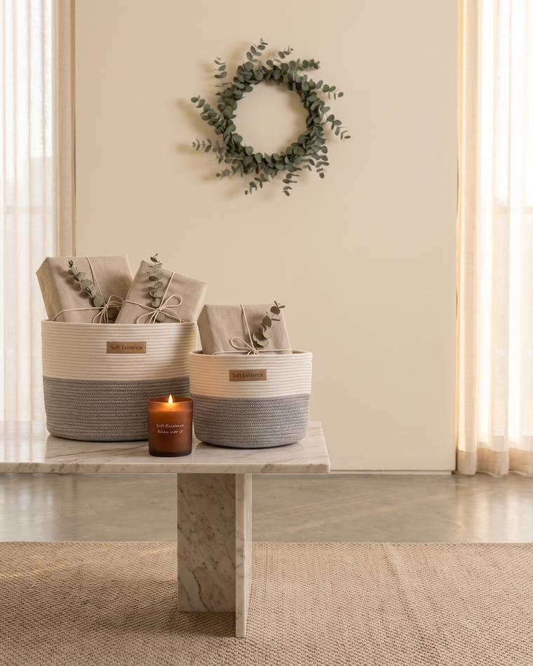 Woven cotton baskets holding linen-wrapped parcels beside a lit amber soy candle and a eucalyptus wreath for a quiet Christmas