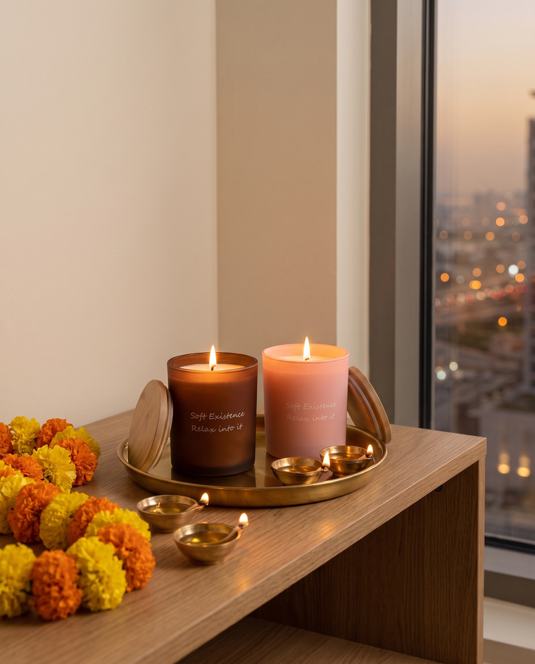 Two lit soy candles in amber and blush glass among brass diyas and a marigold garland on an oak console for Diwali
