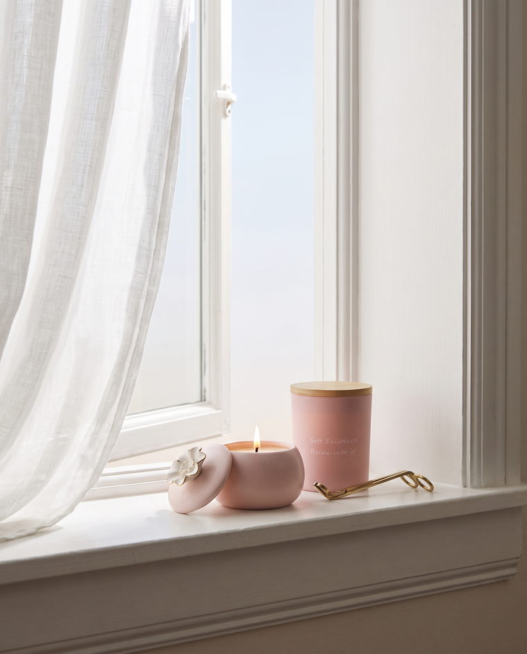 Blush ceramic soy candle burning with a small clean flame on a white windowsill beside an open window, a lidded frosted-glass candle and brass wick trimmers next to it
