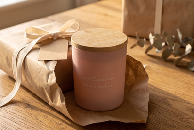 Blush frosted-glass soy candle with a bamboo lid sitting in half-unwrapped kraft paper with a cream ribbon and blank tag on an oak table, a second wrapped parcel behind