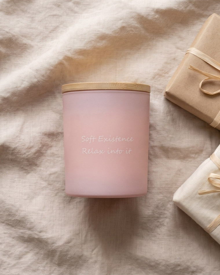 Blush pink frosted soy candle with bamboo lid beside kraft and linen wrapped gifts on beige linen