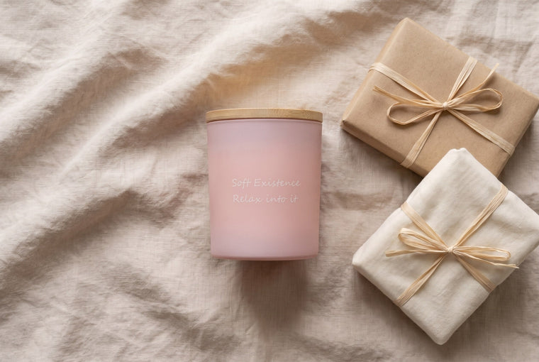 Blush pink frosted soy candle with bamboo lid beside kraft and linen wrapped gifts on beige linen